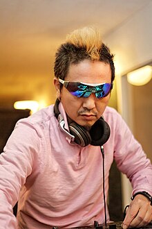 A Japanese man with mirrorshades indoors, a blonde mohawk and headphones around his neck.
