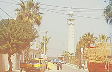 Street in Beni Hassen, Monastir, Tunisia, with palm trees, a mosque's minaret, vehicles, and a hay cart on a sunny day. Power lines and buildings are visible.