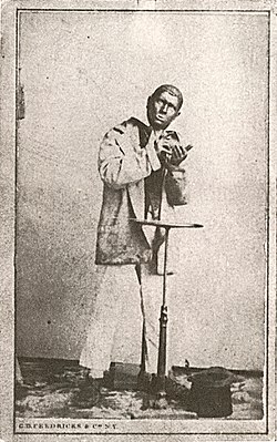 Photo of a man in blackface dressed in nondescript clothes behind a small table with a top hat at his feet.