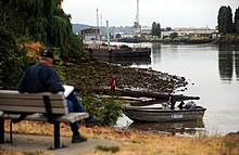 A photograph of a park scene, with a man standing on the shore talking to a man in a small boat, a large barge and warehouses in the background, and a man on a bench reading in the foreground.