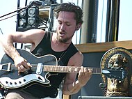 Butler is sitting strumming the strings with his right hand while his left hand is on the fretboard. His hair is relatively short and he is bearded. A microphone is above and to his right with other stage equipment behind his right shoulder. To his left is an oriental statue.