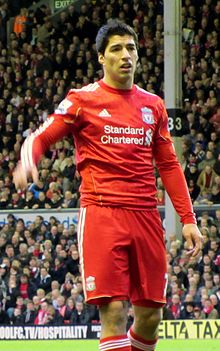 Luis Suárez – wearing a red Liverpool FC jersey with the Standard Chartered sponsor logo at the front centre and shorts with a number 7 partially obscured on the left-leg side and the club crest on the right – lifts his hand with his mouth partly opened.