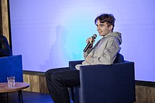 Side view of a smiling Michał Matczak, as he is participating in a panel during a conference