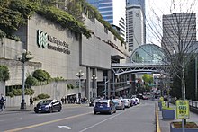 Looking down a street with a queue of vehicles in a left-turn lane. A large concrete wall with the Washington State Convention Center logo is to their left, ahead of an arched bridge over the street.