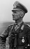 Black-and-white portrait of a man in semi profile wearing a peaked cap, military uniform with an Iron Cross and binoculars suspended from his neck.
