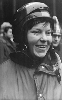A smiling brunette woman wearing a winter jacket and a full-face helmet with a lifted visor.