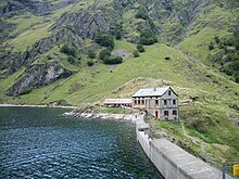 Photographie couleur d'un barrage en béton, retenant un lac. À l'arrière-plan, un refuge et des montagnes.