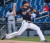 A man in white pants and a navy blue baseball jersey with "Nationals" written across the front throws a pitch.