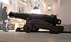 A large black cannon mounted on a wooden frame, located on an inner deck of a ship