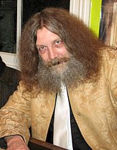 An older caucasian male with long, thick hair and matching beard, sits facing the camera.
