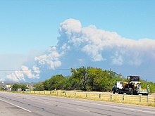 Photograph of a large brown/gray smoke plume emanating from the horizon on a clear afternoon