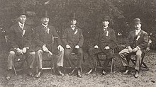 Black and white group photo of five men seated, wearing full-length dress jackets and suits