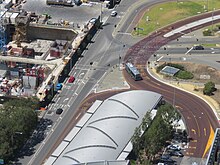 View from a skyscraper of a curved roof which fully covers the station. Next to the station is a busway which enters and exits Elizabeth Quay bus station. East of the station are buildings being constructed at Elizabeth Quay.
