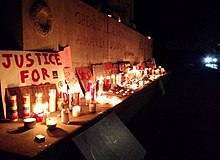 George Floyd–related and Black Lives Matter–themed protest signs and lit candles placed on the base of the Astoria Park War Memorial; the visible portion of the memorial's inscription reads: "GREATER LOVE HATH NO MAN THAN THIS THAT A MAN LAY DOWN HIS LIFE FOR HIS FRIENDS ERECTED 1926 BY THE PEOPLE OF LONG ISLAND CITY" In the background the glaring headlights of a police cruiser.