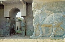 View of a grey stone wall and archway, with the statues of three lamassu (protective deities with wings, the head of a human and the body of a lion or bull).