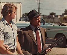 Kirk Douglas (right) holding a gun and John Schneider (left) looking at Douglas, with two light blue trucks in the background, both seen on the set of the movie Eddie Macon's Run during its filming in 1983