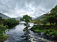 Zusammenfluss mehrerer kleiner Flüsse zu einem Bachlauf, die Ufer sind grün bewachsen mit einigen kleinen Bäumen; in der Ferne sind Berge zu sehen.