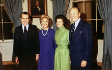 Two women are flanked by two men in suits, standing in a room of the White House.