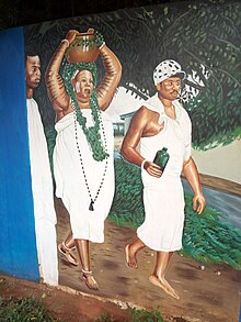 A woman wearing a white cloth carries a pot while clad in green vines walks alongside two men wearing white while also pooring libation