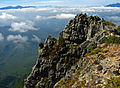 Photo couleur d'une falaise rocheuse. L'arrière-plan est composé d'une nappe de nuages au-dessus d'un massif montagneux boisé.