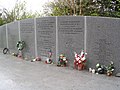 Photo centrée sur le mur en pierre commémoratif. De nombreux bouquets de fleurs et couronnes sont présents à son pied, sur le sol en pierre et il comporte des inscriptions avec les noms des passagers et des phrases en l'honneur des victimes, de leurs proches et premiers secours.