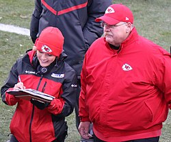 A reporter in a KCTV-branded jacket and writing in a notepad talking to Andy Reid on a football field