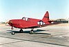 A bright reddish-orange colored aircraft is parked on a concrete ramp with hangars in the background, United States insignia prominent on its side.