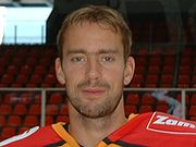 An ice hockey players face and shoulders. He has short brown hair and is not wearing a helmet.