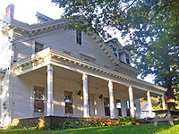 A white wooden house with full=length porch viewed from slightly below at a three-quarter angle lit by early evening sun from the left and sheltered by a large tree on the right