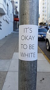 A large white sticker with the slogan on it, wrapped around a lamp post adjacent to an urban sidewalk