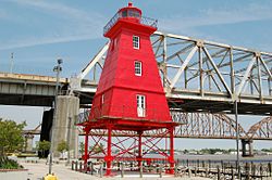 A bright red lighthouse next to a waterfront promenade with multiple bridges in the background.