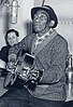 An elderly Mississippi John Hurt sits on a table in front of a suspended microphone, holding a guitar. Another man can be seen in the background.