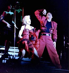 Madonna wearing a magenta jacket and black pants singing to a microphone held in her right hand