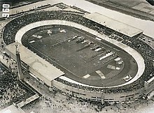 Black and white aerial view of a one-tier stadium surrounding a track and field oval