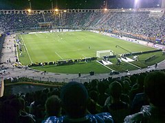 Uma partida de futebol sendo realizada em um estádio com pista de atletismo cheio, vista de trás de um dos gols; é noite e o campo está iluminado por refletores.