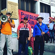 Guatemalan protestors with megaphone standing in front of building and large red sign