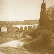 Photographie en sépia représentant le viaduc dans le fond, une petite maison à gauche, un chemin au centre et un cyprès à droite.