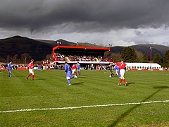 A football match at a football ground. A main stand is visible with hills in the background