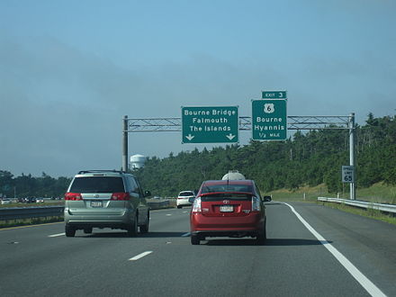 Ground-level view of three lanes of a divided expressway; two large green exit signs are visible in the distance, and the road is surrounded by dense forests.