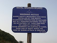 A photograph of a rectangular sign attached to a wooden post. White text on a blue background reads "Natal sharks board. Warning notice. Shark safety measures have been installed at this beach to enhance protection of bathers but all persons entering the sea do so entirely at their own risk. No liability accepted. Avoid swimming or surfing at dawn, dusk and at night when the risk of shark attack is greater. By order."