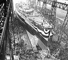 A large warship, still missing most of its superstructure, sits in a dry dock, awaiting its launch. The ship is draped in a large banner and surrounded by crowds of spectators; a huge gantry towers over the ship.