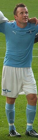 A dark haired man stands with his arms around the shoulders of two teammates, each of whom is out of shot. The subject wears a light blue shirt, white shorts and light blue socks.