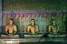 Three stone statues depicting the Buddha sit in meditative repose. In the background, the wall is adorned with paintings.