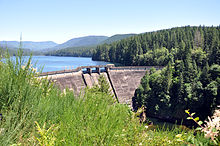 A large concrete dam impounds a lake surrounded by tree-covered hills.