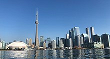 A spear-like tower is between a white-domed structure and small buildings on the left, and increasingly taller buildings to the right. In the foreground is a lake, with a few visible buoys, and the background is a deep blue sky with a few clouds near the horizon.