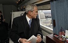 George W. Bush wearing a suit, tie, scarf, and overcoat while looking out the window from a skyward vehicle.