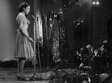 Photographie en noir et blanc, sur une scène, avec sur la gauche, une jeune fille en robe, chantant devant un micro, en bas, sur le coin droit, divers spectateurs.