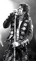 Black and white picture of a man with black hair in a ponytail singing to a microphone; he's wearing a black jacket with buckles, and black pants with straps.