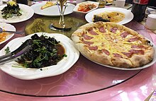 Seven dishes, including a pizza with a slice of meat on each slice, served on a round table.