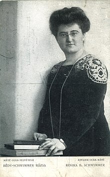 A black and white photograph of a standing woman with her hands placed on three books lying on a table.